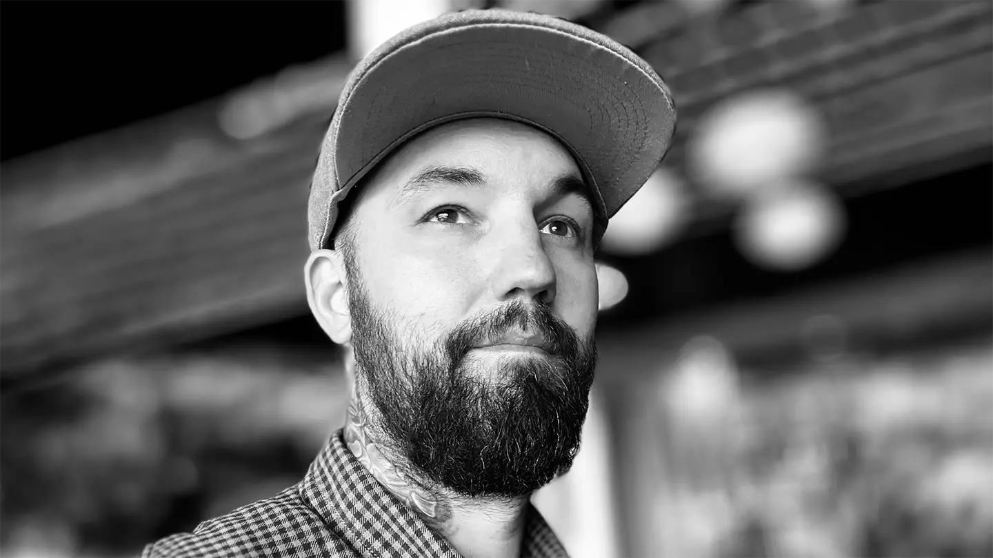 Black and white portrait of tattoo artist with beard and neck tattoos inside studio setting.