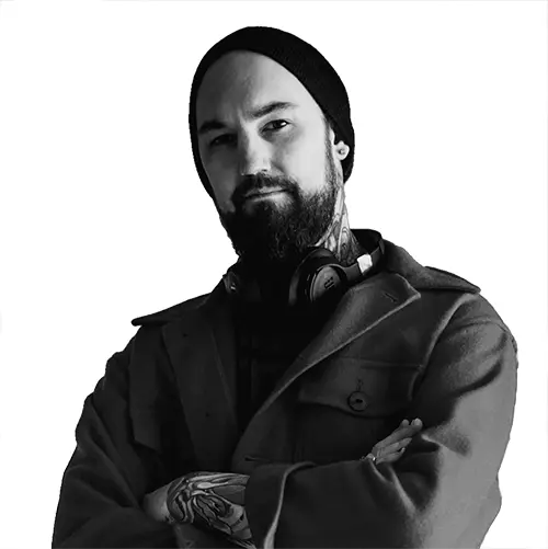 Black and white portrait of tattoo artist standing with arms crossed in front of large mural artwork in studio.